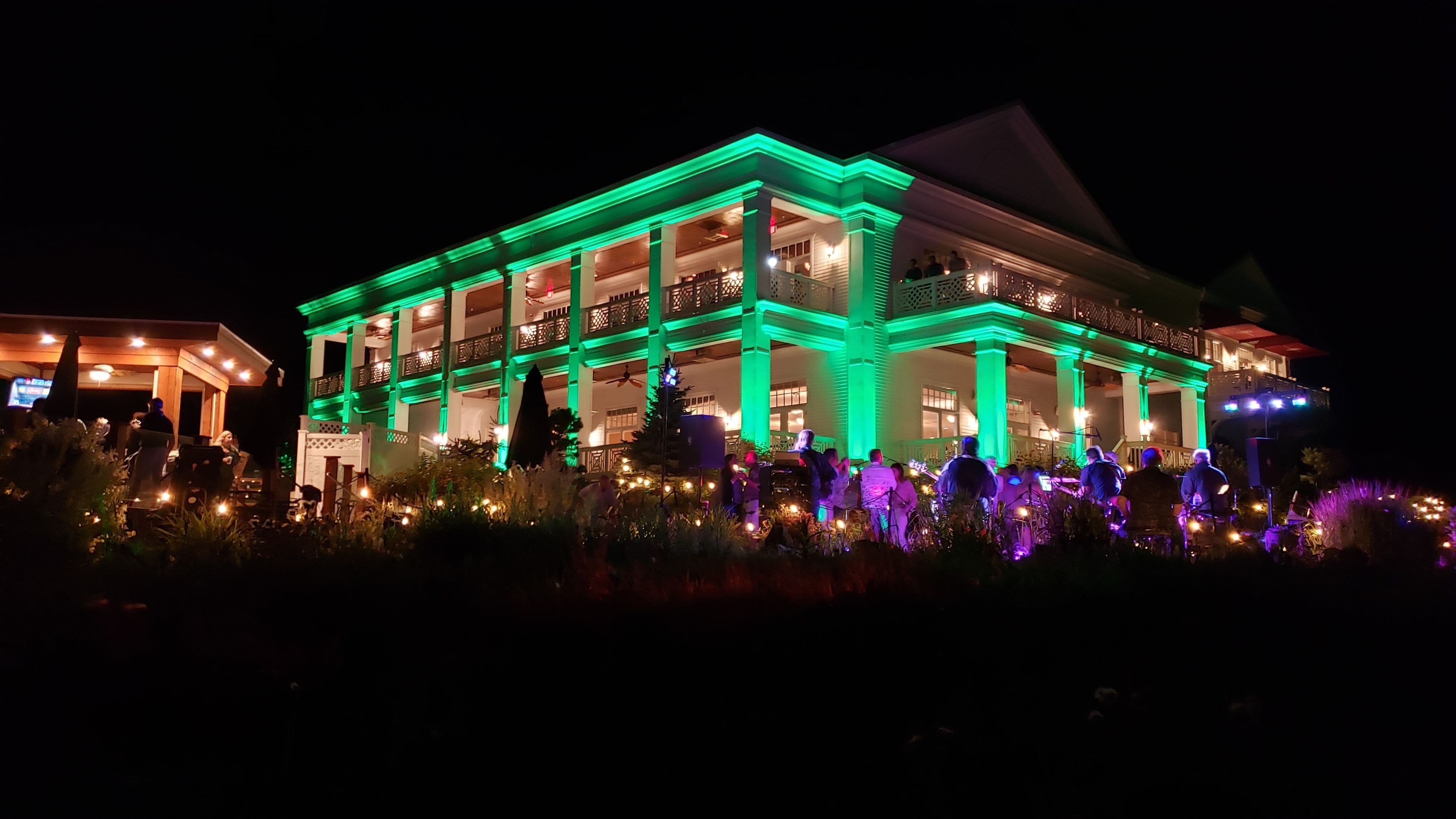 a band plays on the patio of the Northland Country Club with the building lit in green with the gardens lit by bistro.