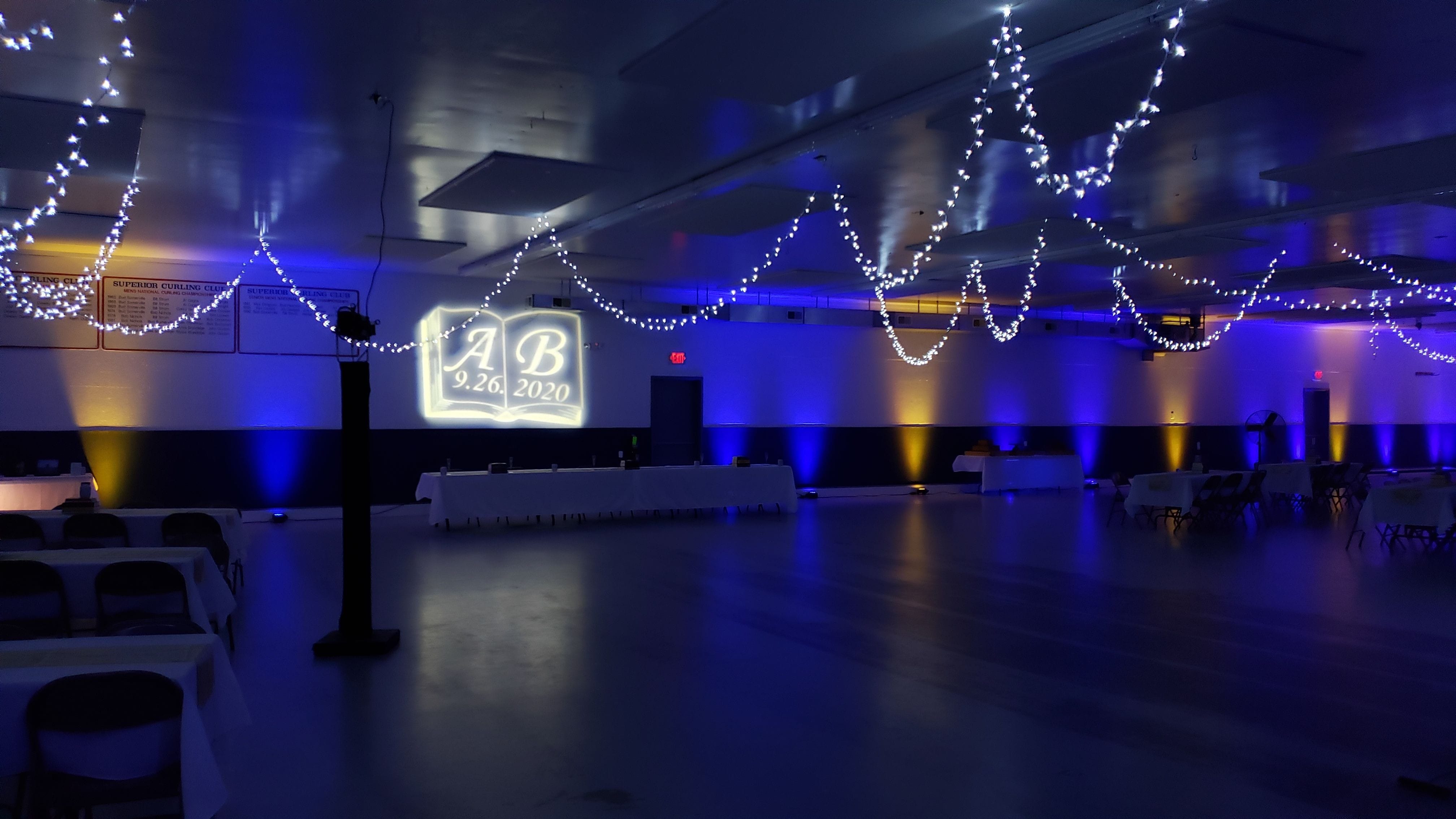 A storybook wedding lighting at the Superior Curling Club. Up lighting in blue and yellow witha wedding monogram of initials in a book.