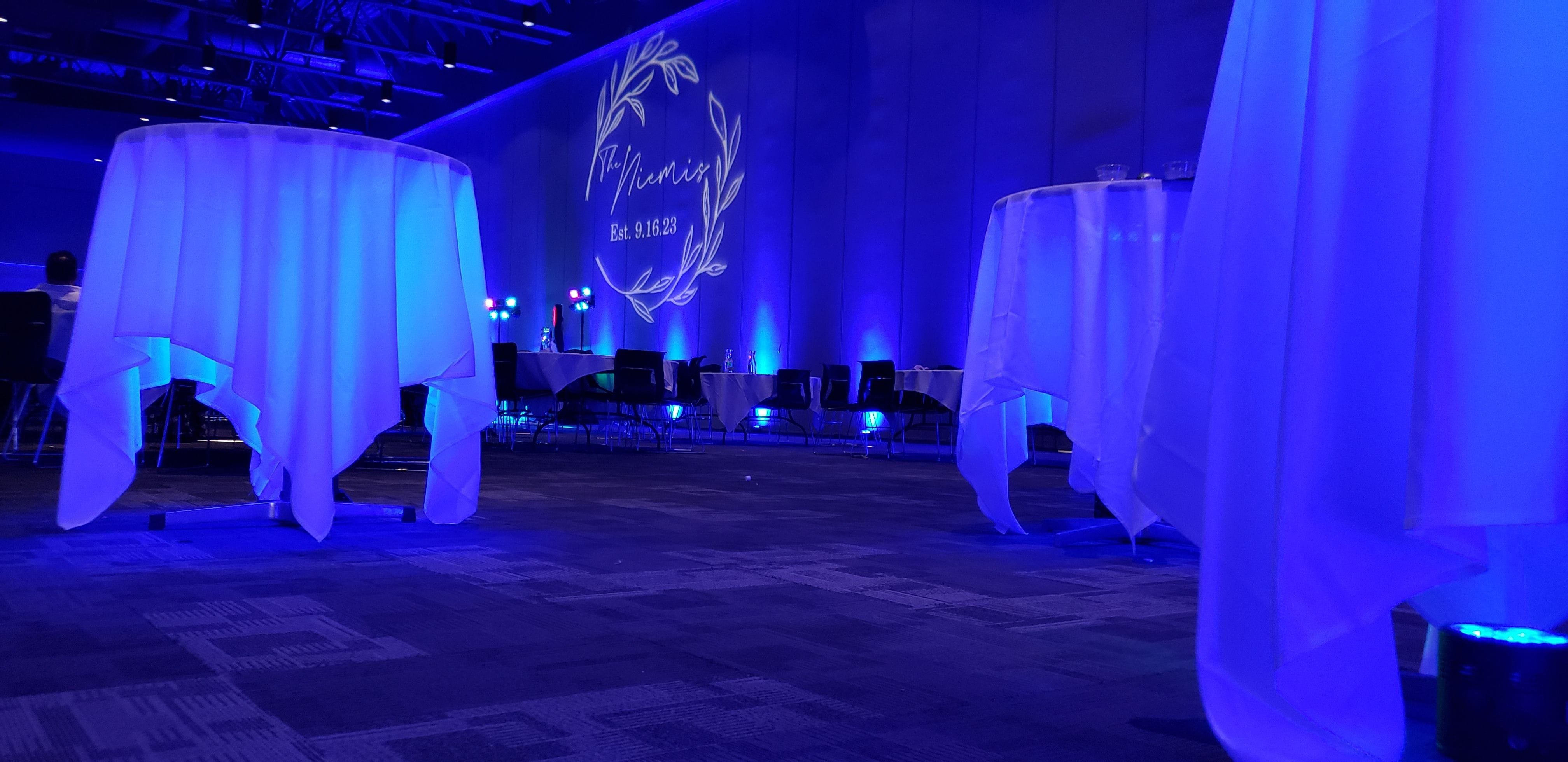 battery lights under cocktail tables to make them glow in blue up lighting with a wedding monogram in the background.